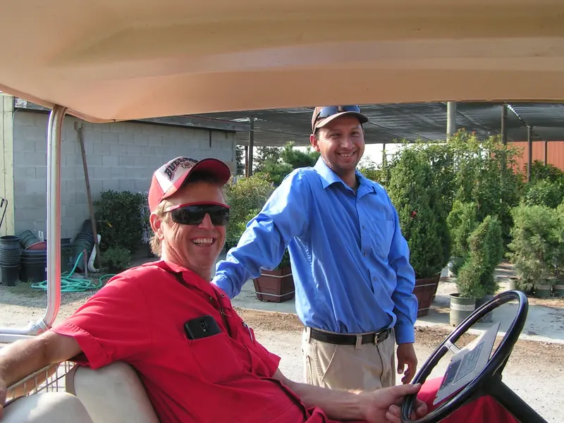 Two men laughing beside a golf cart in the Peters yard, one in a red work shirt at the wheel, container evergreens and a shade house behind them.
